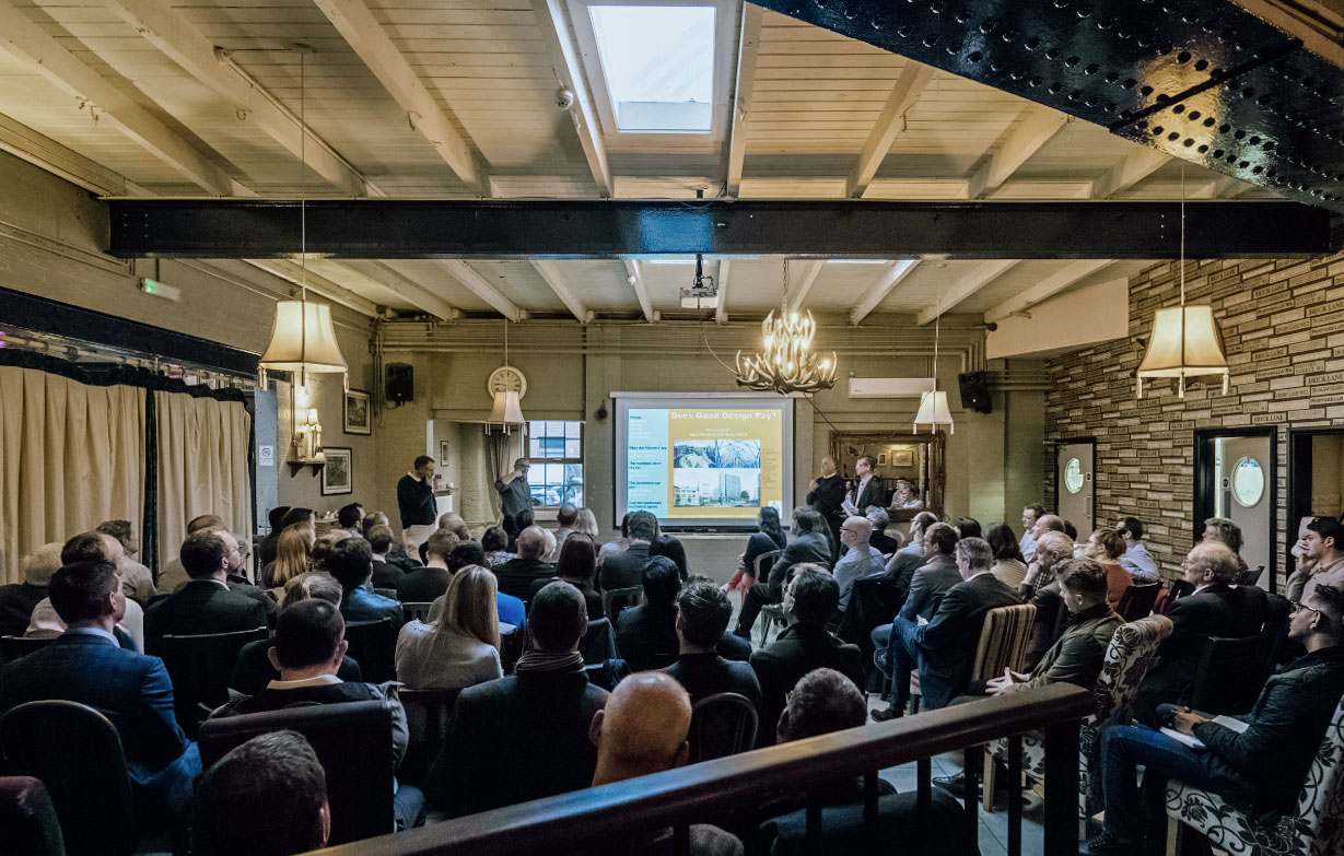 Photograph of man speaking at the front with people sitting infront of him watching him speak and viewing the presentation slides